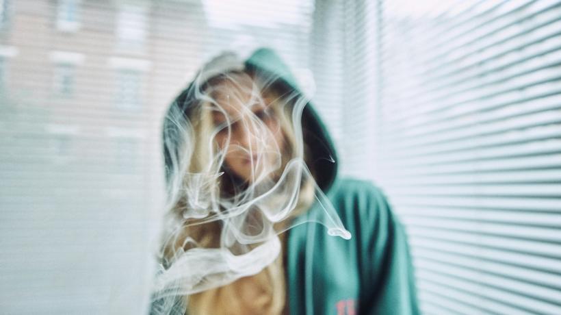 Eine Frau mit langen blonden Haaren und Kapuzenpulli steht vor halbgeschlossenen Jalousien am Fenster. Vor ihrem Gesicht qualmt Rauch.