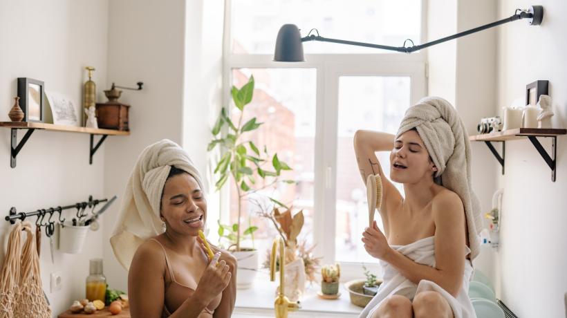 Zwei jungen Frauen sitzen in Handtücher gewickelt im Bad und singen mit Bürsten als Mikrofonersatz.
