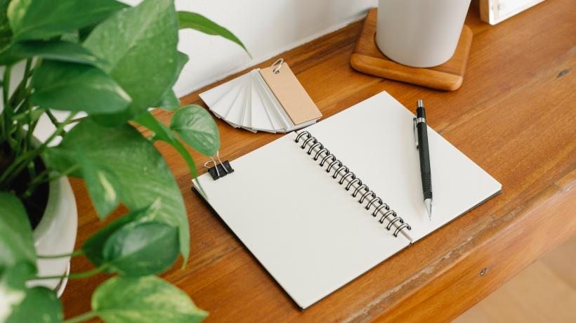 Ein aufgeschlagenes Notizbuch, eine Tasse, ein Stift und eine Pflanze auf einem Holztisch