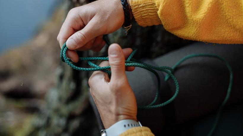 Man sieht die Hände einer älteren Person in gelbem Pullover, die eine grüne Kordel in den Händen hält und einen Knoten entwirrt.
