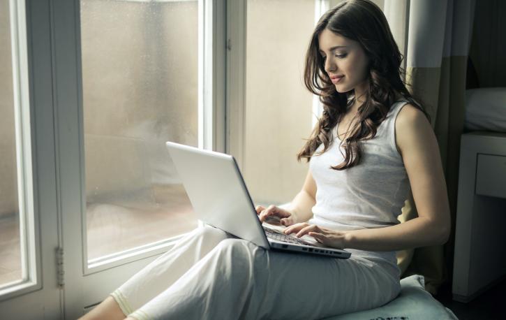 Frau mit Laptop auf den Knien sitzt auf einer Fensterbank