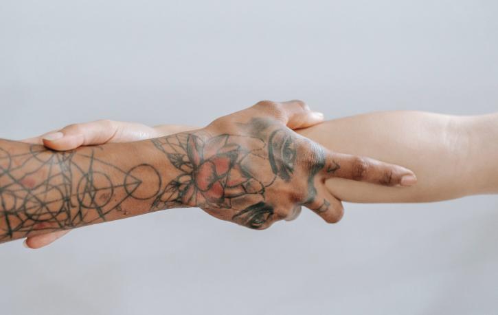 Die Arme zweier Menschen, die sich an den Unterarmen umfassen. Der linke Arm hat Narben, die mit Tattoos übermalt wurden.