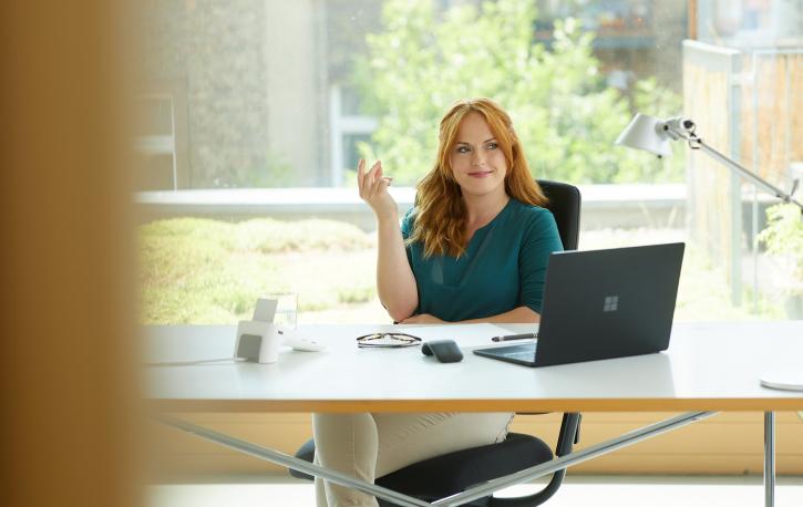 Eine junge Frau sitzt an einem Bürotisch am Laptop und lächelt selbstbewusst.