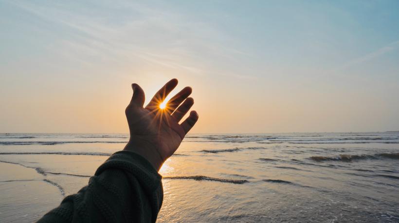Hand vor Meer im Hintergrund, Sonnenstrahlen zwischen den Fingern sichtbar