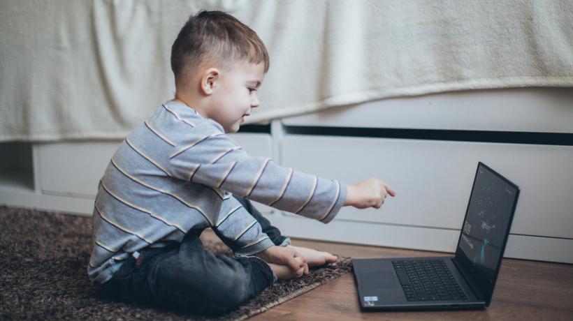 Kleiner Junge sitzt auf dem Teppichboden vor einem aufgeklappten Laptop, auf den er mit dem Finger zeigt.
