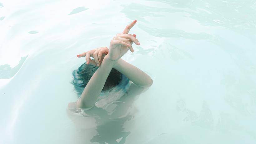 Frau unter Wasser bis zur Stirn. Eine Hand aus dem Wasser heraus in die Höhe gestreckt.