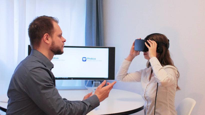 Virtuelle Expositionstherapie kann deinen Patient*innen bei Panik oder Phobie helfen. (Foto: Phobius) Mann und Kind sitzen am Tisch. PC im Hintergrund. Kind hat VR-Brille auf. Man erklärt mit den Händen.
