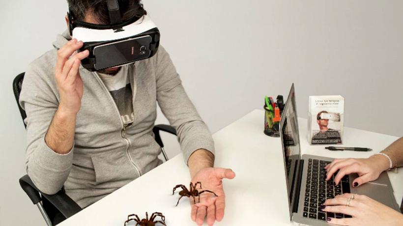 Mann sitzt am Tisch, trägt VR-Brille. Eine Hand liegt offen auf dem Tisch. Zwei große Spinnen laufen über Hand und Tisch.