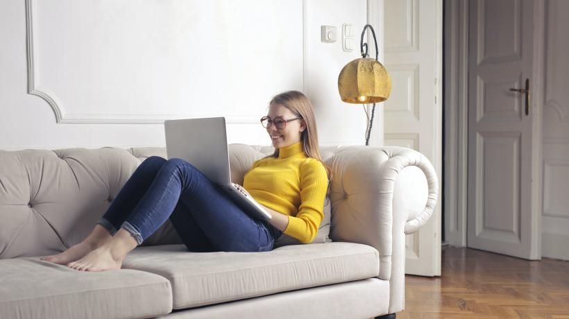 Eine junge blonde Frau liegt lächelnd mit einem Laptop auf dem Schoß auf der Couch.