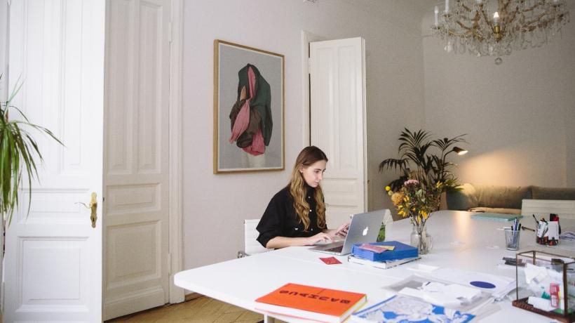 (Foto: Polina Zimmermann – Pexels.com) Altbauzimmer, im Vordergrund ein Tisch mit Pflanze und Papieren. Eine junge Frau sitzt vor einem aufgeklappten Laptop.