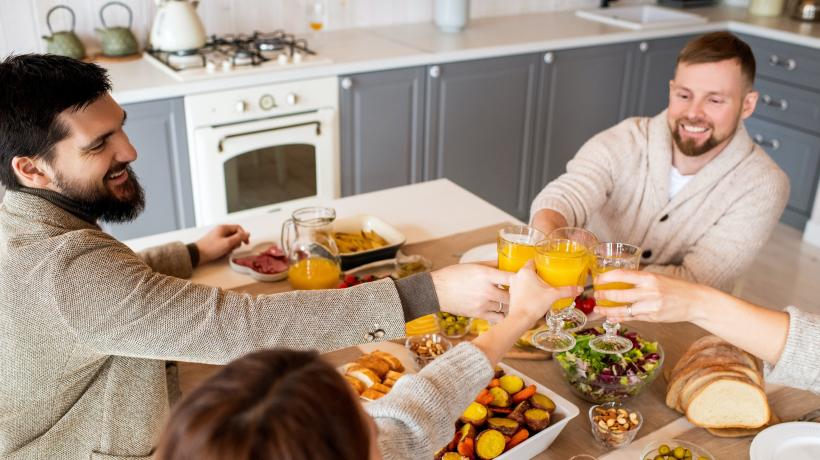 (Alexy Almond – Pexels.com) Vier Freunde sitzen am Essenstisch und stoßen mit Orangensaft an.