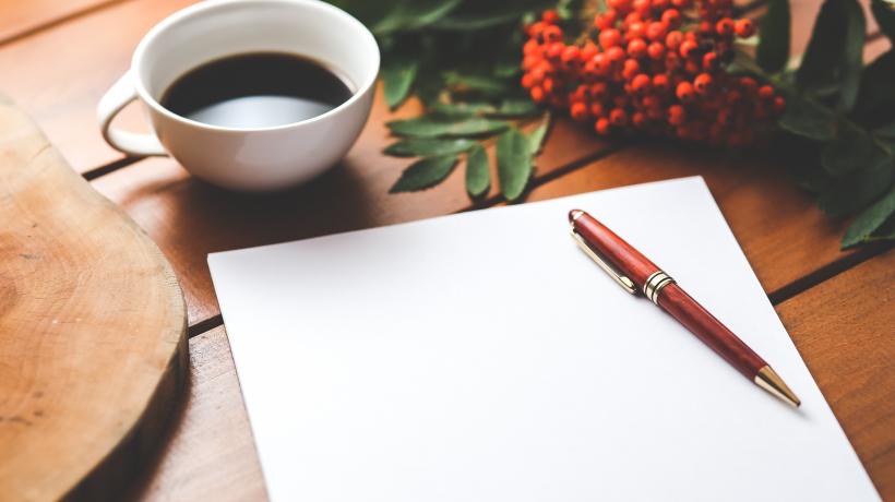 (Foto: Kaboompics – Pexels.com) Auf einem Tisch steht eine Tasse Kaffe und davor liegt ein Schreibblock mit einem Stift.