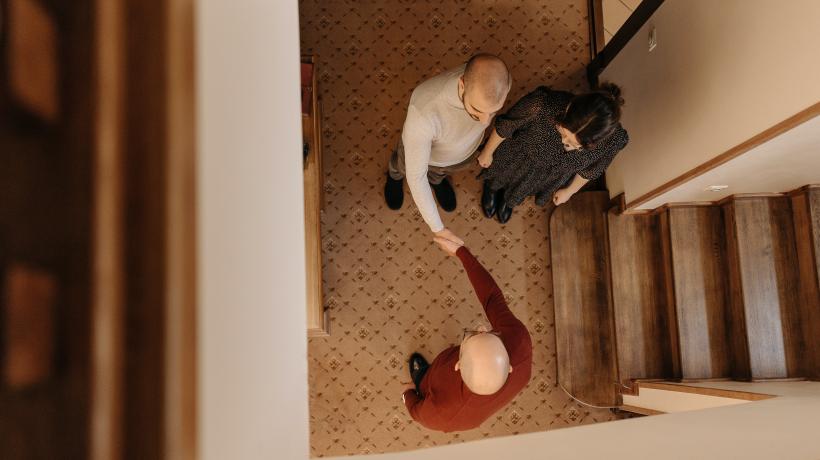 Bild von oben im herunten ins Treppenhaus. Drei Menschen stehen sich gegenüber. Zwei schütteln einander die Hand.