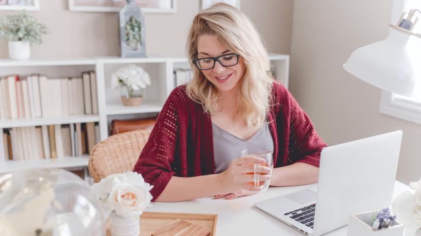 (Foto: Paige Cody – Unsplash.com) Blonde Frau sitzt lächelnd am Schreibtisch vor aufgeklapptem Laptop.