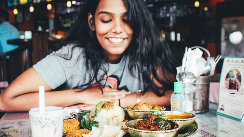 Junge Frau sitzt im Restaurant und lächelt über dass appetitliche indische Essen, das vor ihr auf dem Tisch steht.