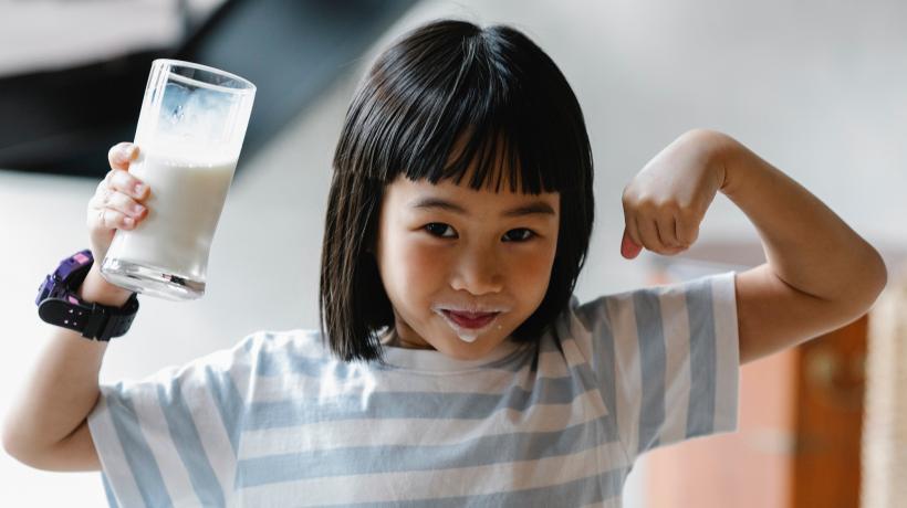 Kleines Mädchen mit einem Milchglas in der Hand und Milchbart spannt die Muskeln an und lächelt in die Kamera.