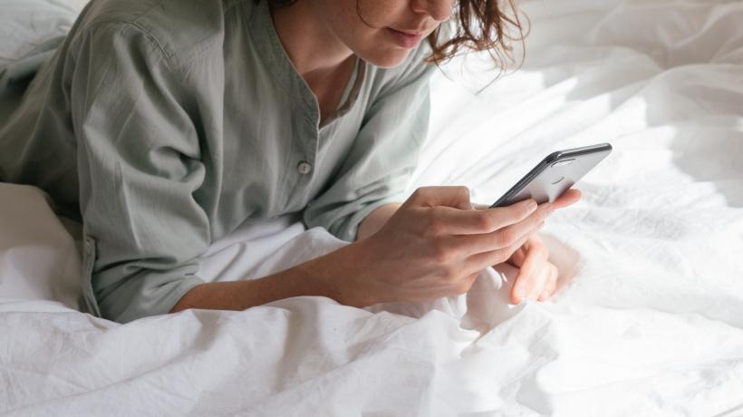 Junge Frau liegt auf dem Bauch auf dem Bett und schaut auf ihr Smartphone.