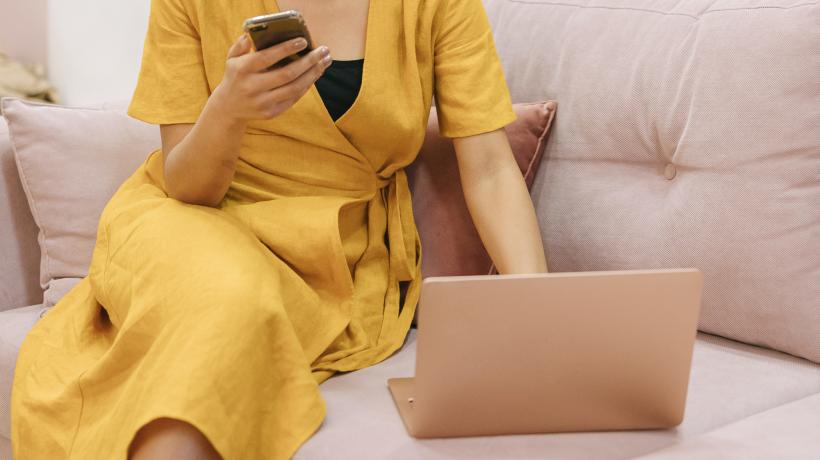 Frau in gelbem Kleid sitzt vor Laptop mit Handy in der Hand.