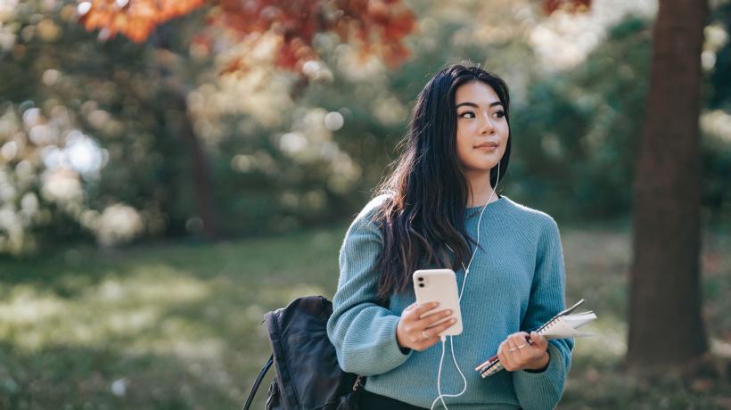 Junge Frau mit Rucksack auf dem Rücken und Unterlagen in der einen, Handy in der anderen Hand läuft durch den Park und hört etwas über Kopfhörer.