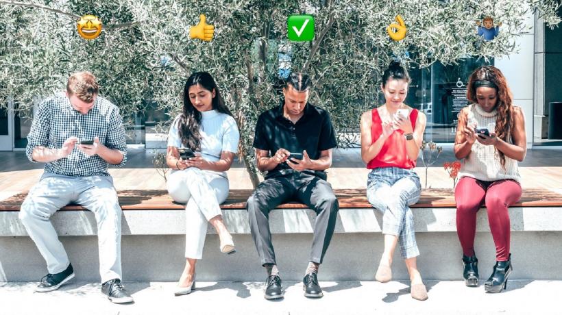 Zwei Männer und drei Frauen sitzen nebeneinander auf einer Bank, tippen auf ihren Smartphones herum. Über ihren Köpfen sind unterschiedliche Emojis abgebildet.