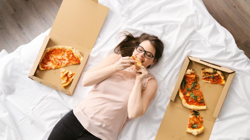 Bild von Cornelia Fiechtl, die auf einem Bett liegt, zwei Pizzakartons neben sich, und genüsslich in ein Stück Pizza beißt.