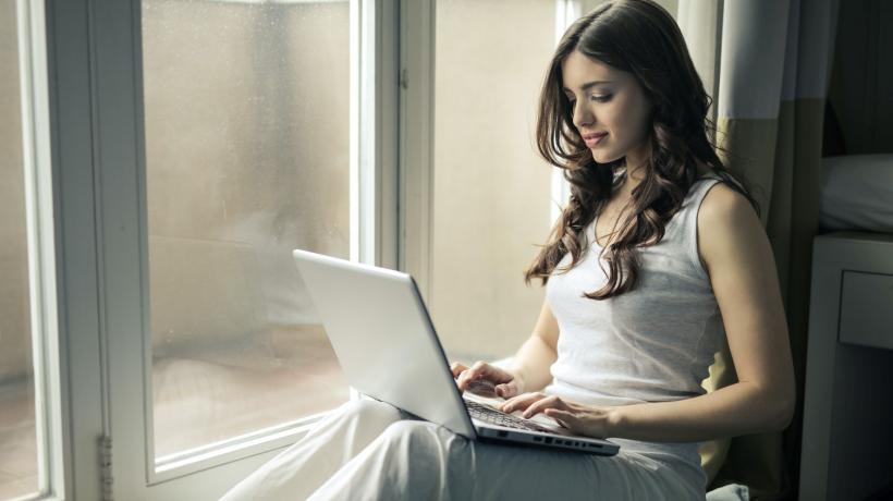 Frau mit Laptop auf den Knien sitzt auf einer Fensterbank