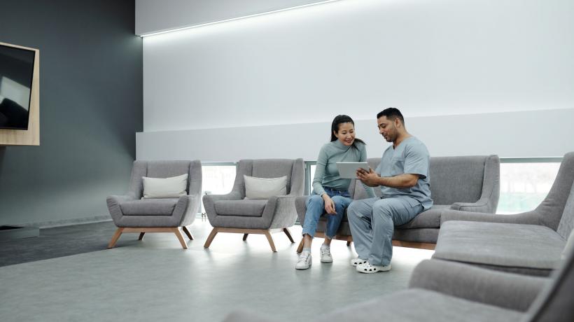 Mann und Frau sitzen auf Sesseln in einem Wartezimmer und schauen auf ein Tablet.
