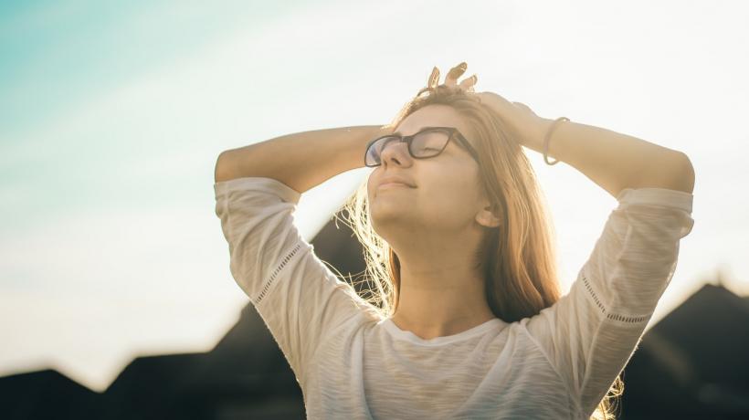 Eine junge Frau mit Brille streckt versunken und mit geschlossenen Augen ihr Gesicht nach oben, die Hände hat sie entspannt hinter ihren Kopf gelegt. Ihr Haar schimmert im Sonnenlicht. Im Hintergrund sind Hügel zu sehen.