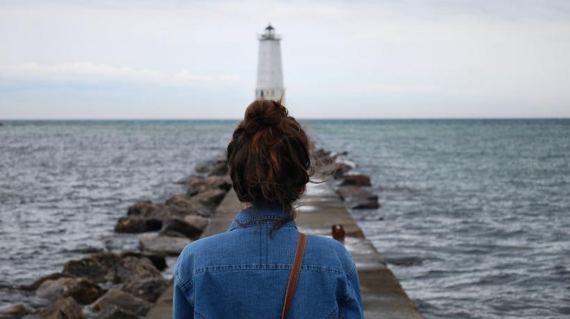 (Foto: Will Wilson – unsplash.com) Eine Frau steht mit dem Rücken zur Kamera an einem Steg und blickt in Richtung eines Leuchtturm, der am Ende des Stegs zu sehen ist. Links und rechts des Stegs ist Wasser.