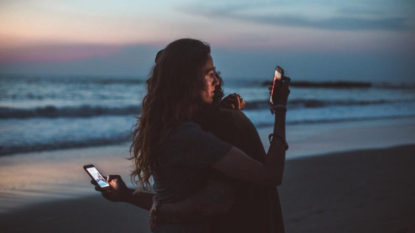 Ein Paar steht am Strand und umarmt sich, aber dabei schauen beide auf ihr jeweiliges Handy.