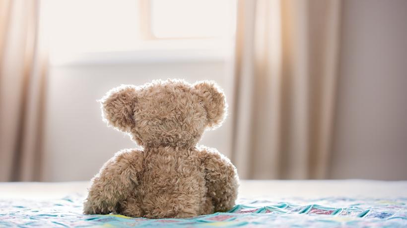Ein Teddybär von hinten, auf einem Bett sitzend und aus dem Fenster schauend.