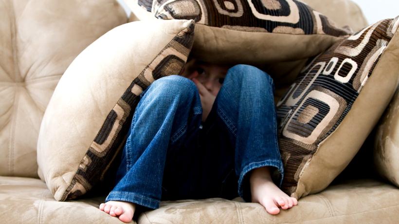 Ein Junge hat sich auf der Couch unter drei Kissen vergraben und schaut ängstlich aus seiner Kissenhöhle hervor.
