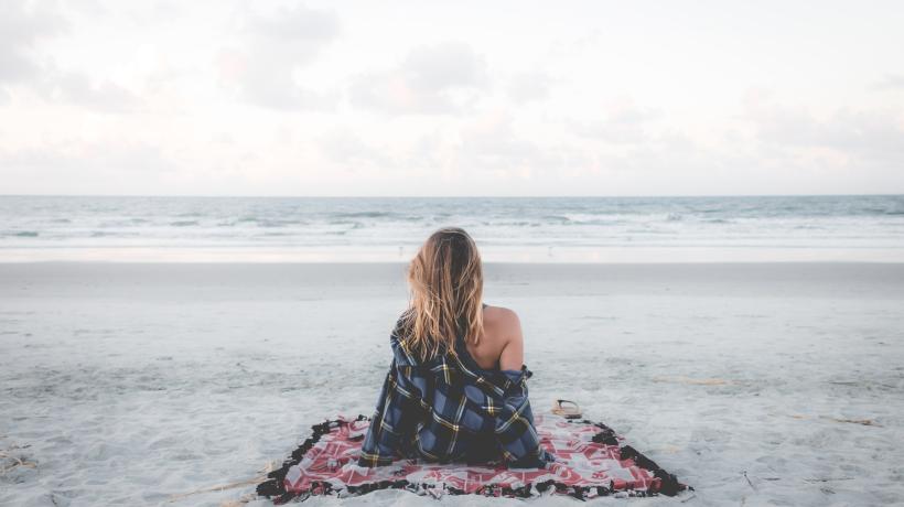Eine Frau sitzt auf einer Decke am Strand und blickt aufs Meer.