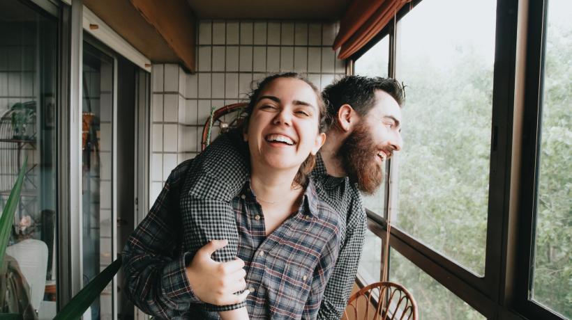 Ein Mann und eine Frau stehen auf einem verglasten Balkon und lachen, dabei hat er von hinten lässig einen Arm um sie geschlungen, den sie mit ihrer Hand festhält.