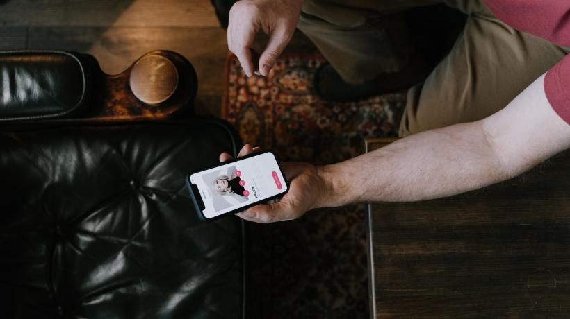 Ein Mann sitzt auf einem Ledersessel und blickt auf sein Handy, auf dessen Bildschirm eine Dating-App zu sehen ist.