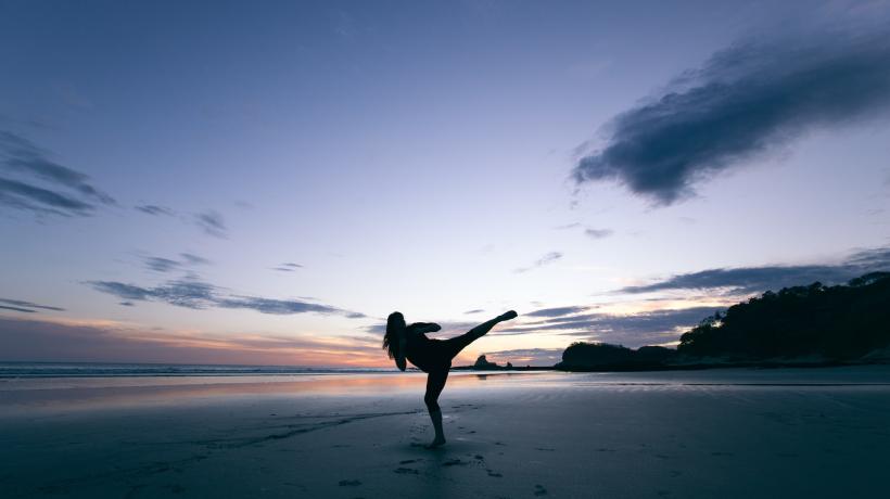 Bei Sonnenuntergang ist die Silhoutte einer Frau zu sehen, die am Strand einen hohen Beinkick aus dem Kampfsportbereich ausführt.