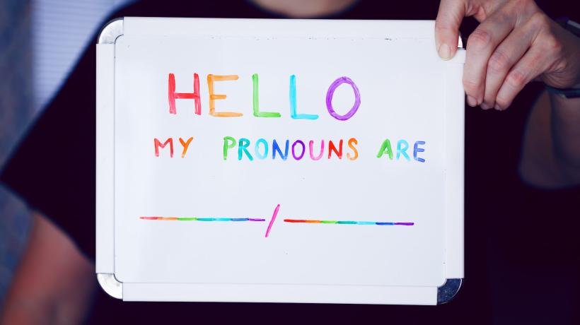 Eine Person hält ein Schild vor sich, auf dem "Hello, my pronouns are..." geschrieben steht.