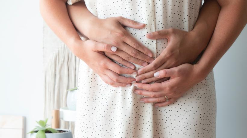 Bildausschnitt, auf dem man sieht, wie eine schwangere Frau die Hände auf ihren Bauch legt, eine zweite Person umarmt sie von hinten und hat ihre Hände ebenfalls auf den schwangeren Bauch gelegt.
