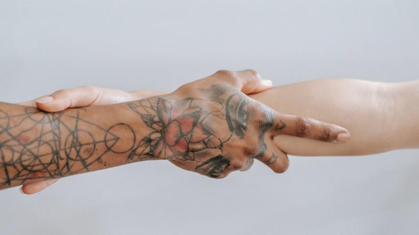 Die Arme zweier Menschen, die sich an den Unterarmen umfassen. Der linke Arm hat Narben, die mit Tattoos übermalt wurden.