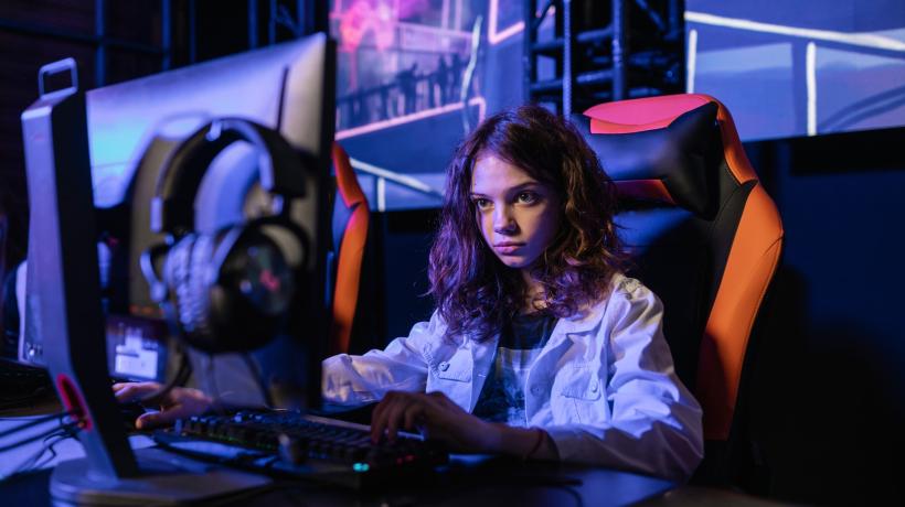 Ein junges Mädchen sitzt in einem dunklen Raum vor einem Computer und starrt auf den Bildschirm. Die rechte Hand liegt auf ihrer Maus, die linke auf der Tastatur.