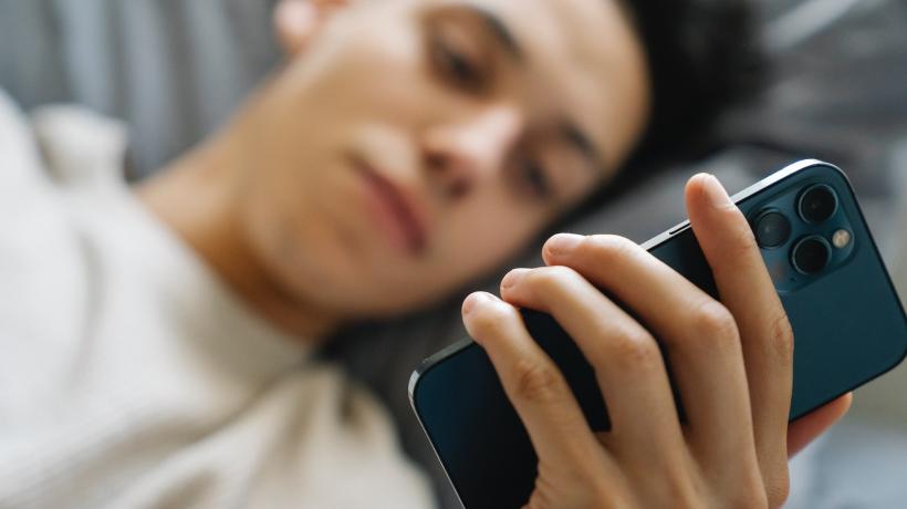Eine junge Person liegt mit dem Kopf auf einem Kissen und hält ihr Handy in der Hand, auf dessen Display sie schaut.