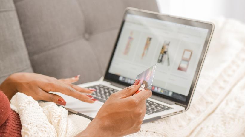 Die Hände einer Person mit rot lackierten Fingernägeln sind zu sehen, mit der linken Hand bedient sie ihr Notebook und surft in einem Onlineshop, in der rechten Hand hält sie eine Kreditkarte.
