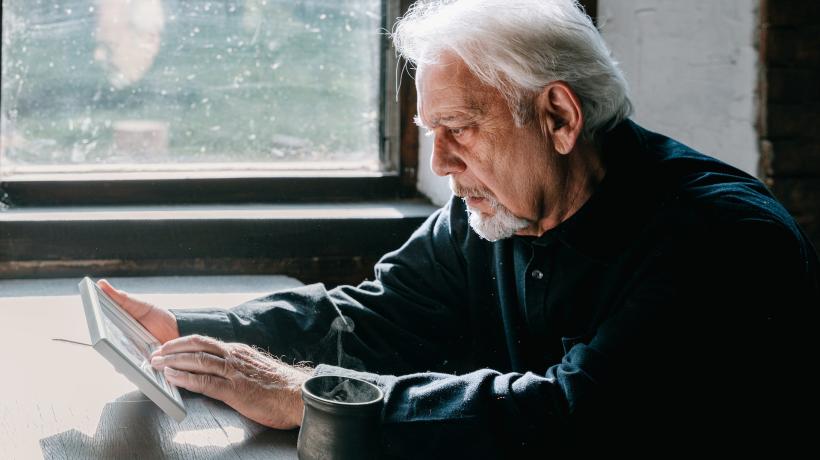 Ein weißhaariger, älterer Mann sitzt am Tisch, blickt traurig auf ein Foto und streicht mit der linken Hand darüber.