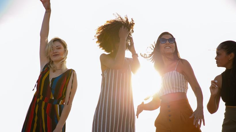 Vier junge Frauen tanzen im Freien, hinter ihnen ist die Sonne zu sehen.