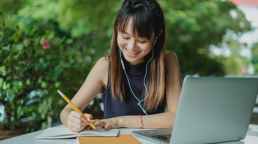 Eine junge Frau sitzt mit Laptop und Arbeitsmaterialien an einem Tisch und schreibt mit einem Stift in ein Heft.