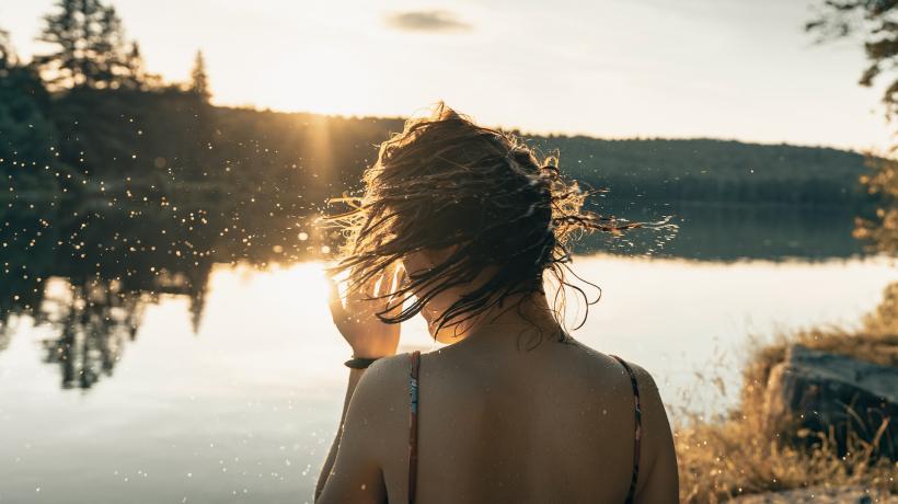 Eine Frau steht im Badeanzug an einem See und schüttelt ihre nassen Haare.