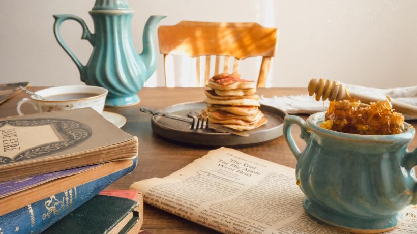 Auf einem Tisch steht ein Teller mit Pancakes, daneben eine Kanne Kaffe, ein Stapel Bücher und eine Zeitung.