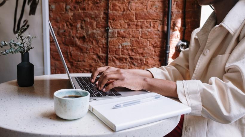 Eine Person arbeit an einem Laptop, neben sich auf dem Tisch ein Notizblock mit Stift sowie eine Tasse Kaffee.