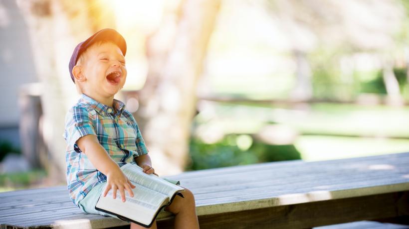 Ein kleiner Junge sitzt mit einem Buch auf den Knien lachend auf einem Tisch und lacht aus vollem Hals.