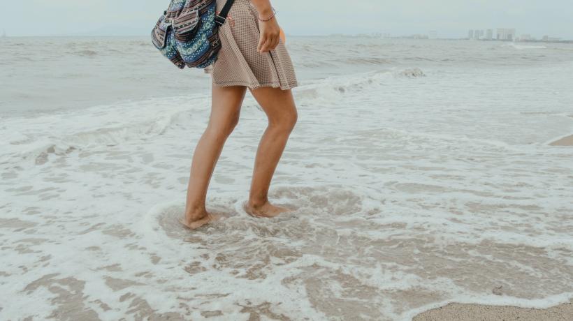Der Unterkörper einer Person ist zu sehen, die mit nackten Füßen durch Wellen am Strand läuft.